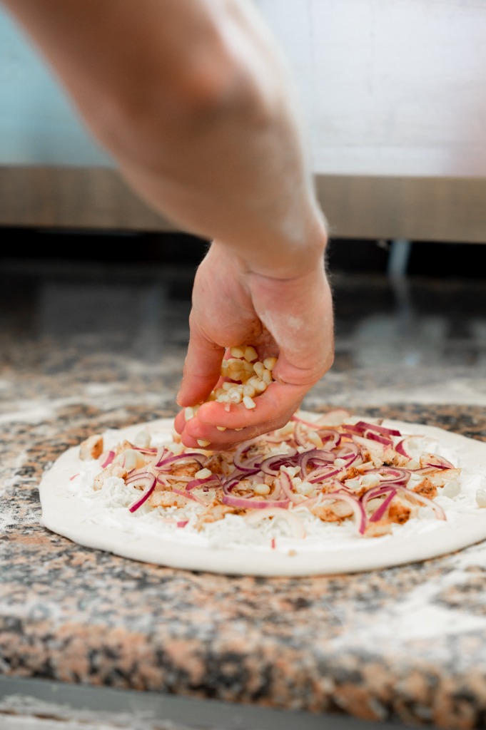 Pizzakészítő morzsolt kukoricát szór a lilahagymás baconös pizzára